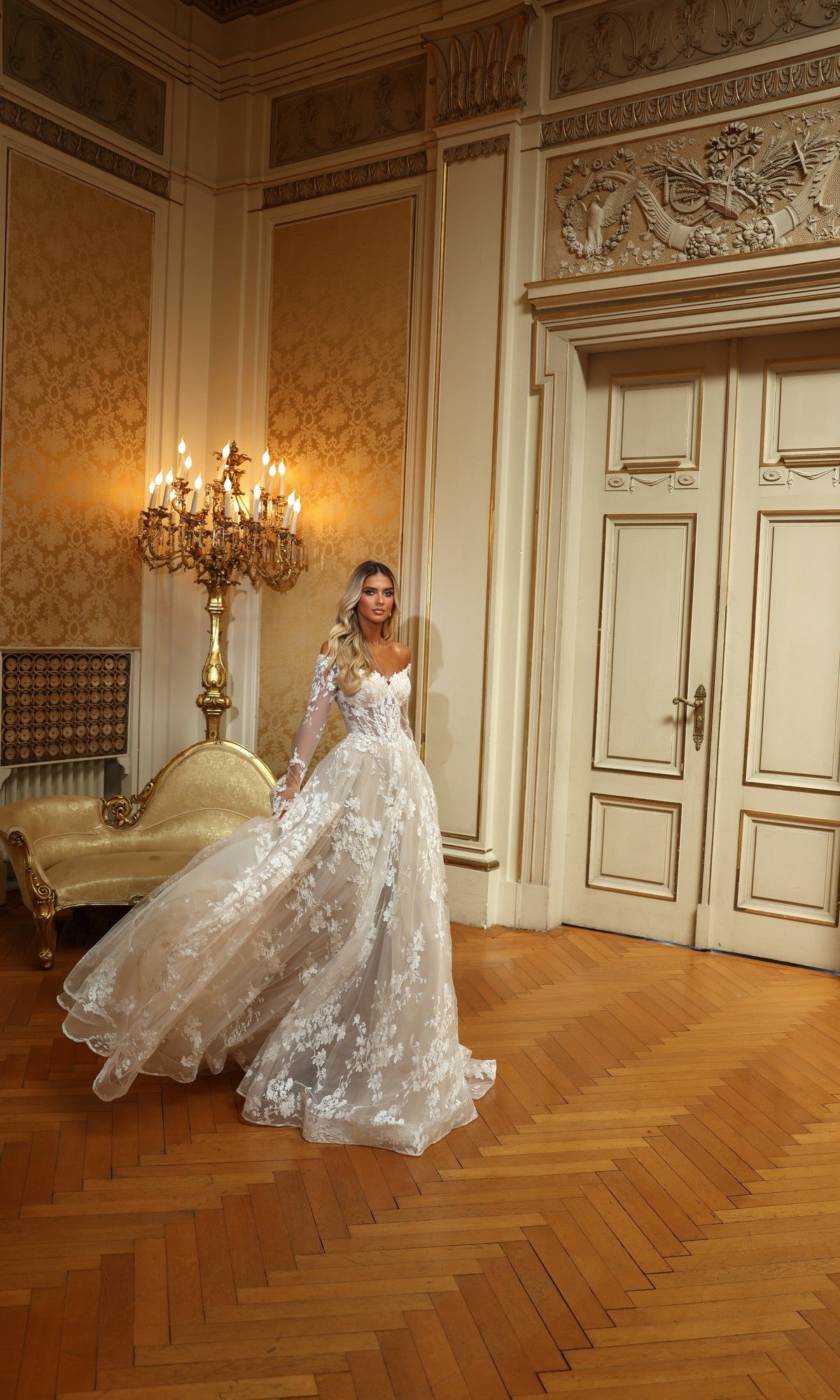 A woman wearing the Alyce 7150 long white dress with lace and ruffle details stands in an ornate room with wooden floors, gold-patterned wallpaper, a candelabra, and a cream-colored settee.